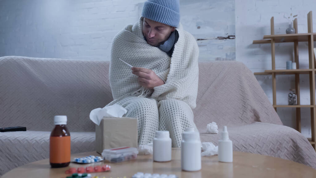 ill man met warme hoed en deken kijken naar thermometer in de buurt van tafel met medicijnen en papieren servetten - Foto, afbeelding