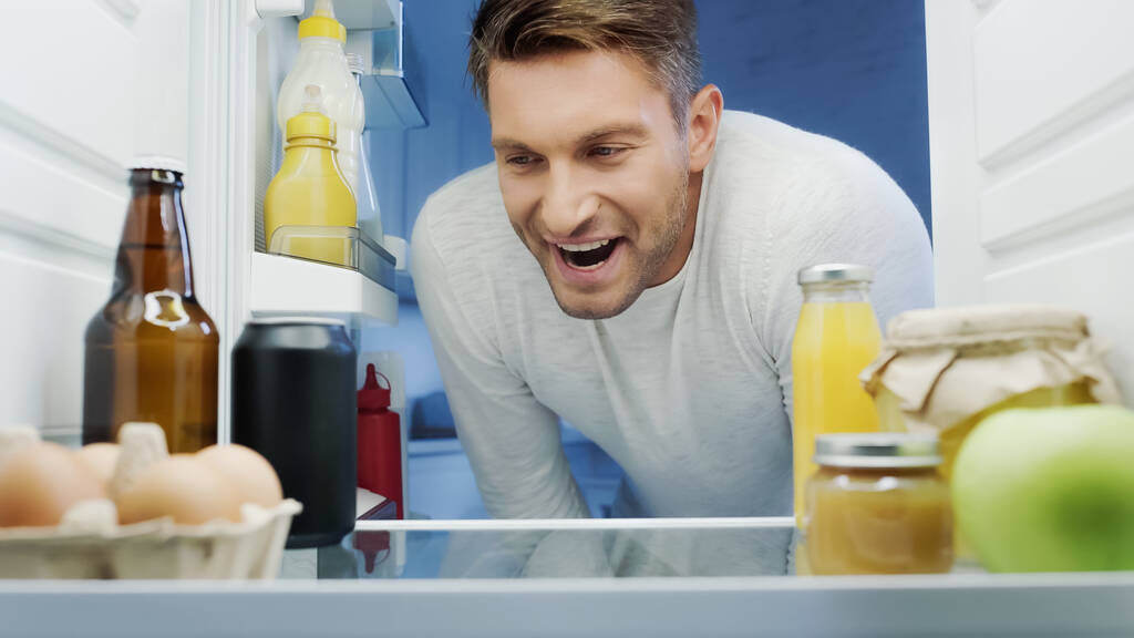 excited man looking into fridge with drinks, eggs, jars and bottles with sauces - Foto, Imagen