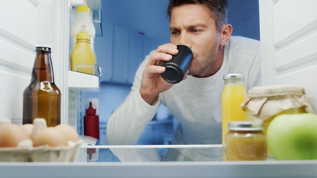 άνθρωπος πίνοντας σόδα από μεταλλικό κουτί κοντά στο ψυγείο με ποτά, αυγά και μπουκάλια με σάλτσες - Φωτογραφία, εικόνα