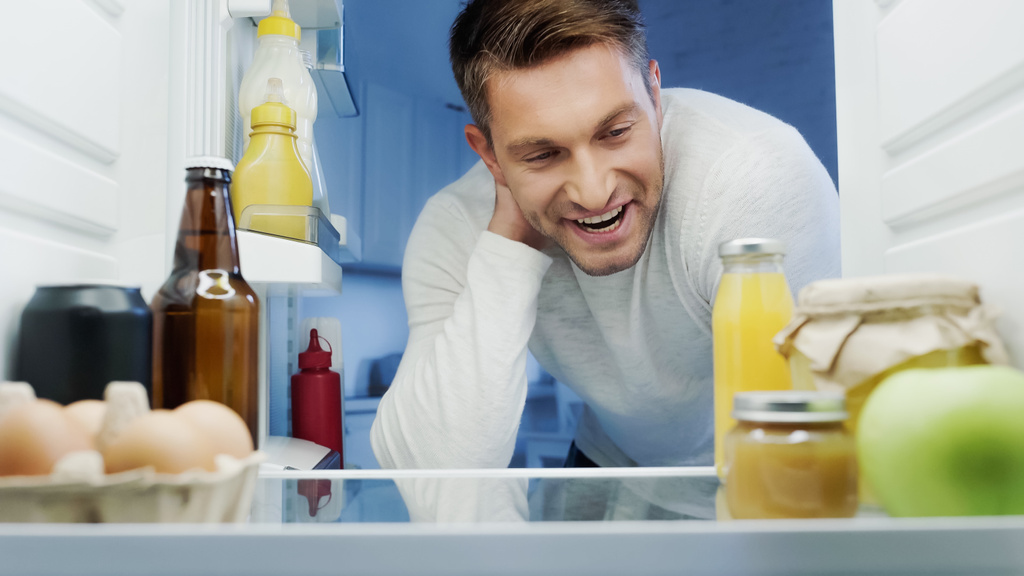 veselý muž hledající do ledničky s čerstvým pomerančovým džusem, pivem, vejci a lahvemi s omáčkou - Fotografie, Obrázek