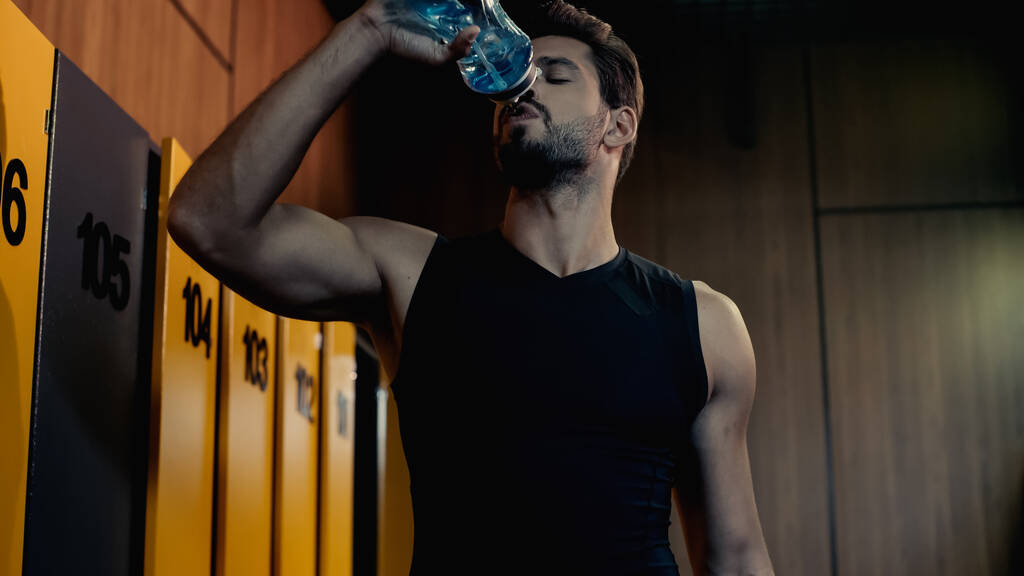 bearded sportsman drinking water from sports bottle in locker room  - Foto, afbeelding