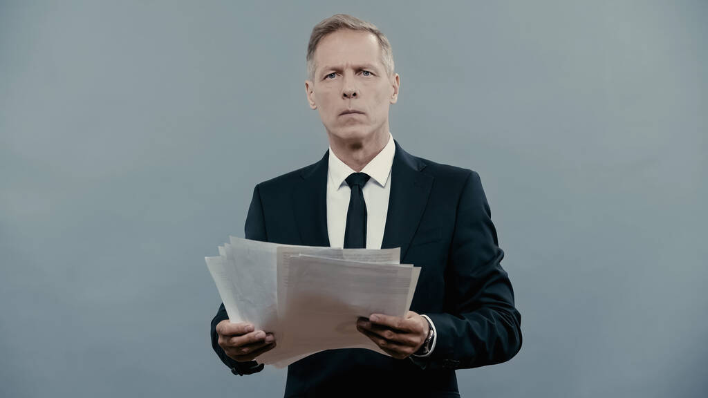 Mature manager in suit holding documents and looking at camera isolated on grey  - Foto, Imagen