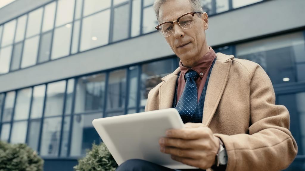 Зрілий бізнесмен в пальто використовує розмитий цифровий планшет на міській вулиці
  - Фото, зображення