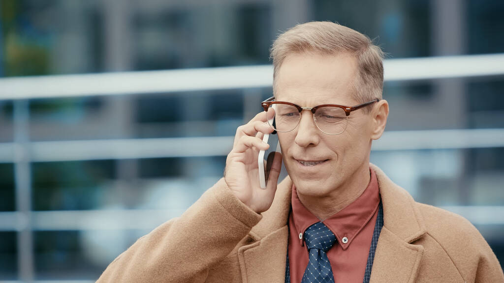 Homme d'affaires souriant en manteau parlant sur un téléphone portable dans une rue urbaine  - Photo, image