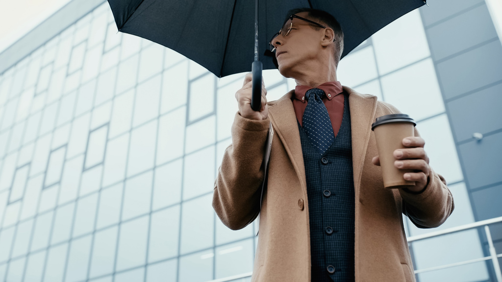 Low angle view of businessman holding coffee to go and umbrella on urban street  - Foto, Imagen