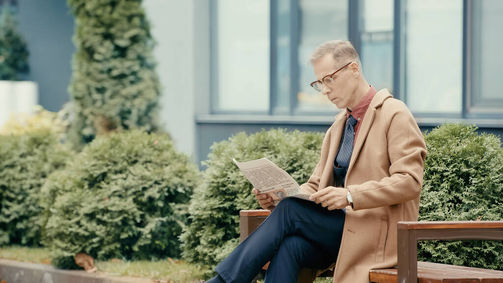 Mature businessman in coat reading newspaper on bench on urban street  - Valokuva, kuva