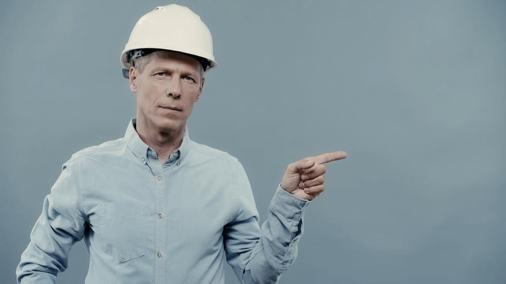 Ingeniero maduro en casco apuntando con el dedo y mirando a la cámara aislada en gris  - Foto, Imagen