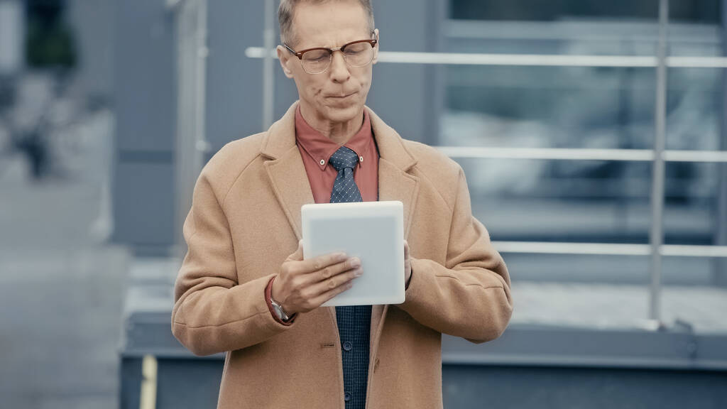 Middle aged businessman in coat using digital tablet on urban street  - Photo, Image