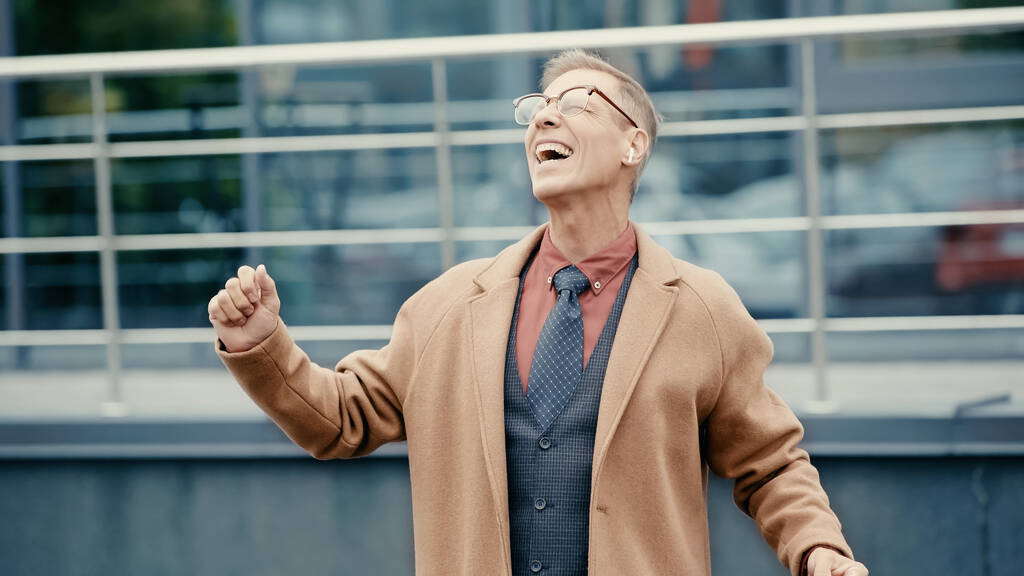 Excited businessman in coat and earphone singing on urban street  - Foto, Imagen
