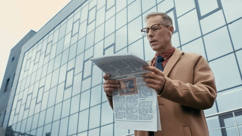 Низький кут зору бізнесмена в пальто і навушники читання газети на міській вулиці
  - Фото, зображення