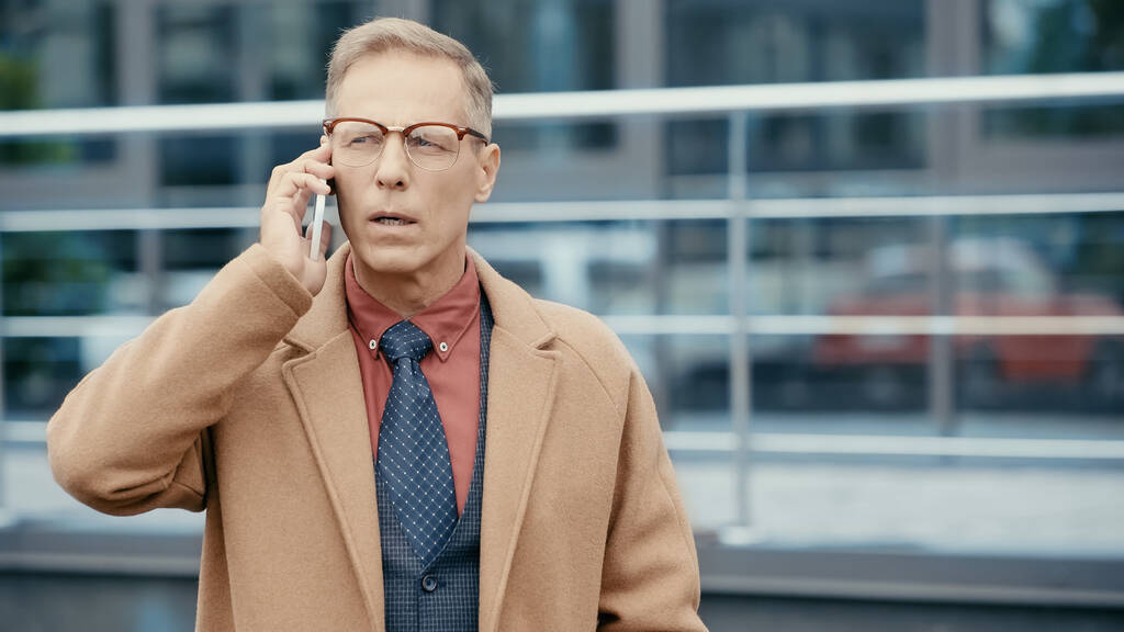 Бізнесмен середнього віку в формальному одязі та окулярах розмовляє на смартфоні на відкритому повітрі
  - Фото, зображення