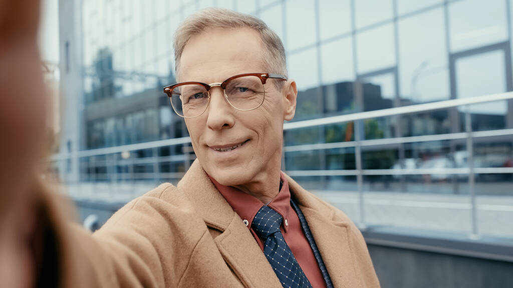 Smiling businessman in coat and eyeglasses looking at camera on urban street  - Foto, afbeelding