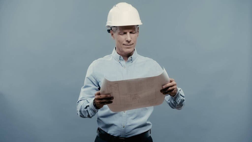 Smiling engineer in hardhat looking at blueprint isolated on grey  - Foto, Imagem