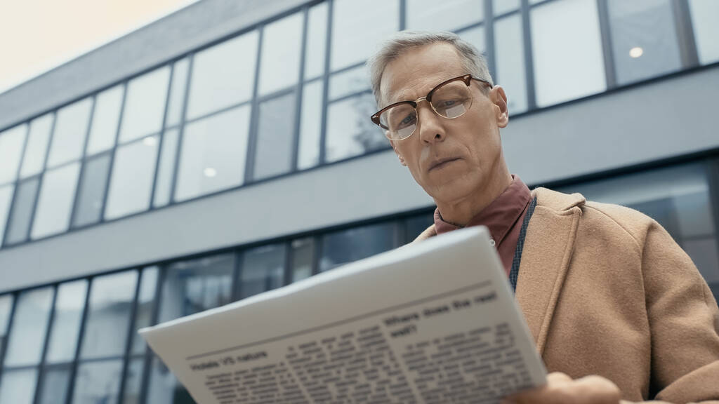 Χαμηλή γωνία άποψη του ώριμου επιχειρηματία σε παλτό και γυαλιά ηλίου ανάγνωση ειδήσεων στο αστικό δρόμο  - Φωτογραφία, εικόνα