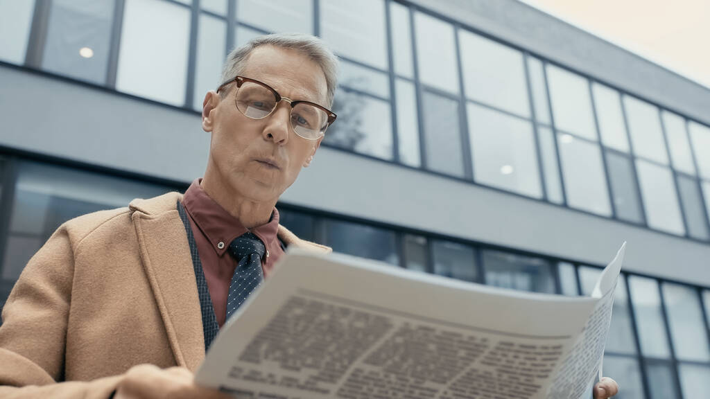 Empresario en abrigo leyendo periódico cerca de difuminado edificio al aire libre  - Foto, imagen