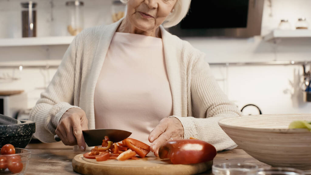 vista recortada de la mujer anciana cortando pimiento en la tabla de cortar en la cocina - Foto, Imagen