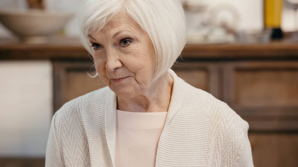 mujer mayor con el pelo gris mirando hacia otro lado en la cocina borrosa - Foto, Imagen