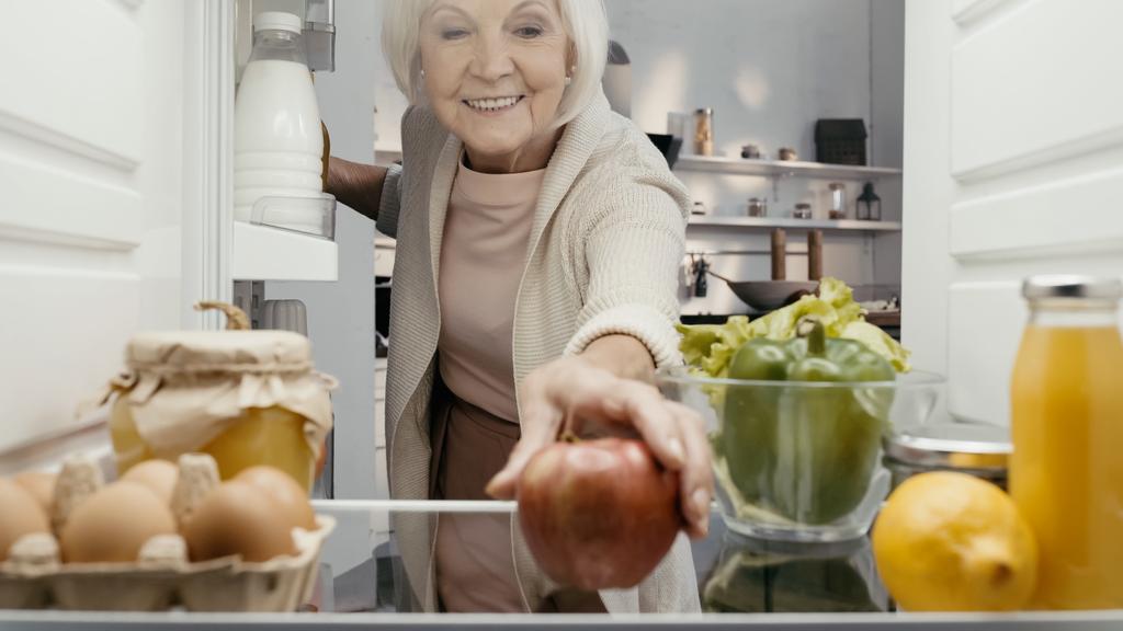 feliz anciana tomando manzana fresca de la nevera con verduras frescas, frutas, bebidas y huevos - Foto, Imagen
