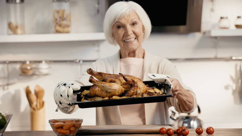 щаслива старша жінка тримає лист печі зі смачною індичкою та картоплею біля свіжих овочів, розмитим фоном
 - Фото, зображення