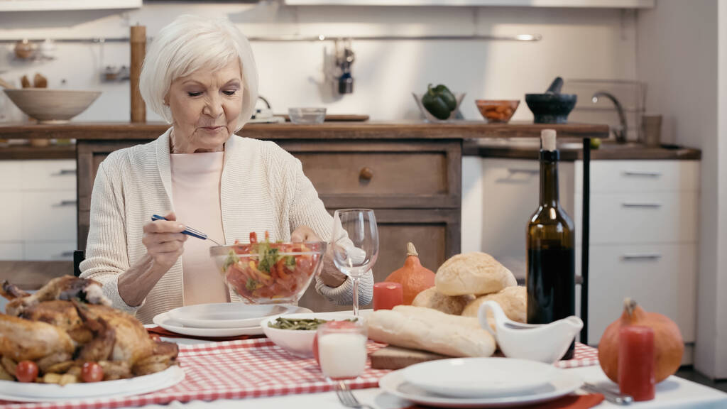 mujer mayor sosteniendo tazón de ensalada de verduras cerca de pavo asado y vino tinto en la mesa - Foto, Imagen