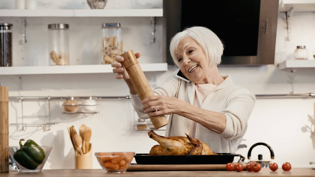 ευτυχισμένη ηλικιωμένη γυναίκα καρυκεύματα ψητό γαλοπούλα, ενώ μιλάμε στο smartphone - Φωτογραφία, εικόνα