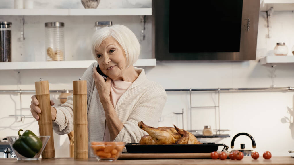 positive senior woman talking on mobile phone near roasted turkey, spice mills and fresh vegetables - Фото, изображение