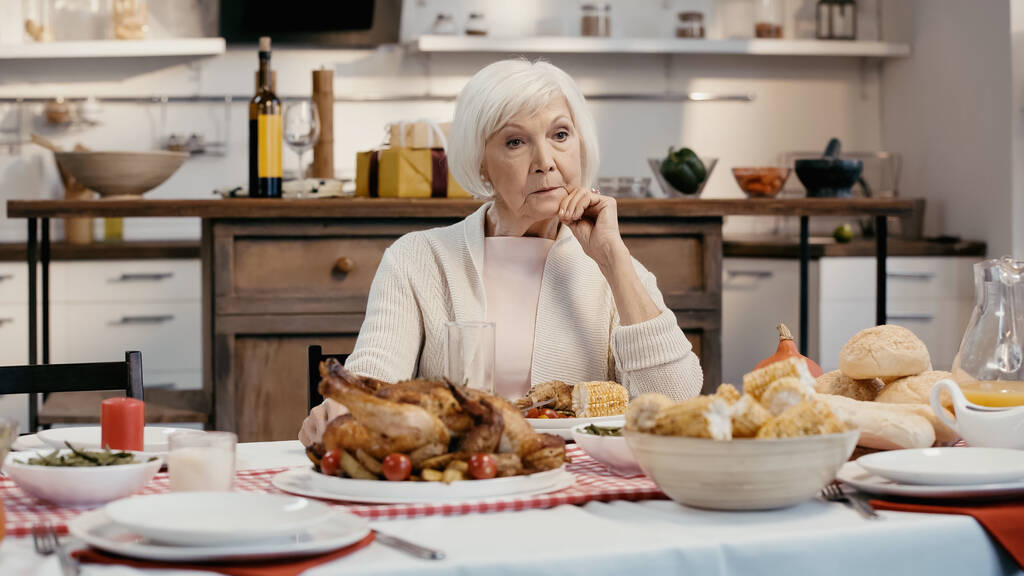 mujer anciana solitaria y pensativa sentada cerca de la cena de acción de gracias servida en la mesa en la cocina - Foto, Imagen