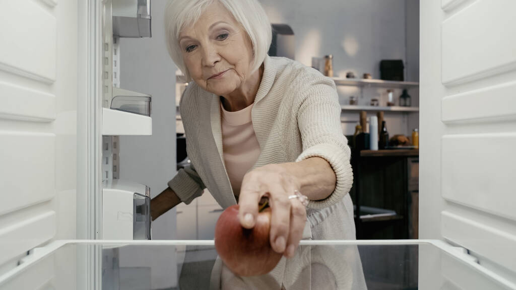 старша жінка бере стигле червоне яблуко з холодильника на кухні
 - Фото, зображення