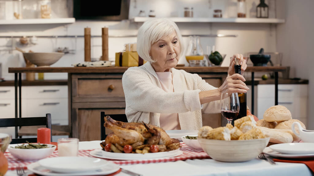 ηλικιωμένη γυναίκα ξεπακετάρει μπουκάλι κόκκινο κρασί κοντά στο ψητό γαλοπούλα, ψητό καλαμπόκι και ψωμάκια - Φωτογραφία, εικόνα