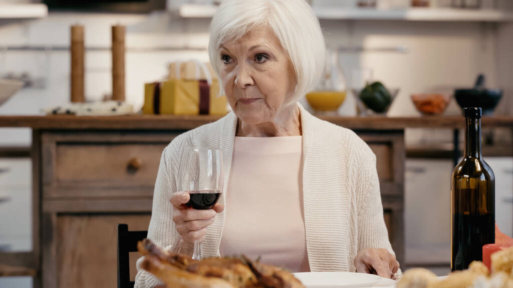 senior woman holding glass of red wine near blurred roasted turkey during thanksgiving dinner - Foto, Imagen
