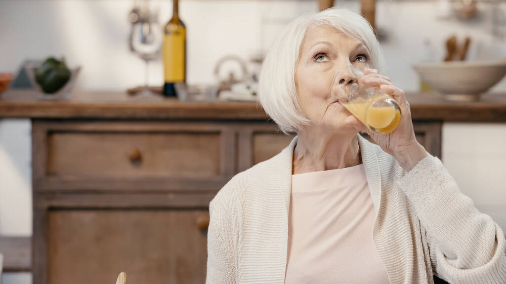 senior woman drinking orange juice in blurred kitchen - Фото, изображение