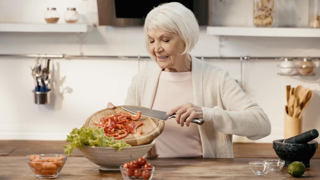 smiling senior woman adding sliced bell pepper to lettuce while preparing vegetable salad - Foto, Imagen