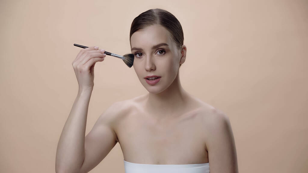 young woman with bare shoulders applying face powder while holding cosmetic brush isolated on beige - Zdjęcie, obraz