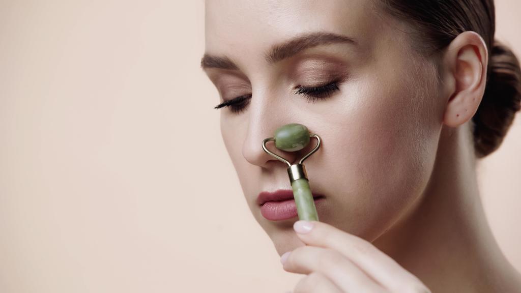young woman using jade roller on nose isolated on beige  - Valokuva, kuva