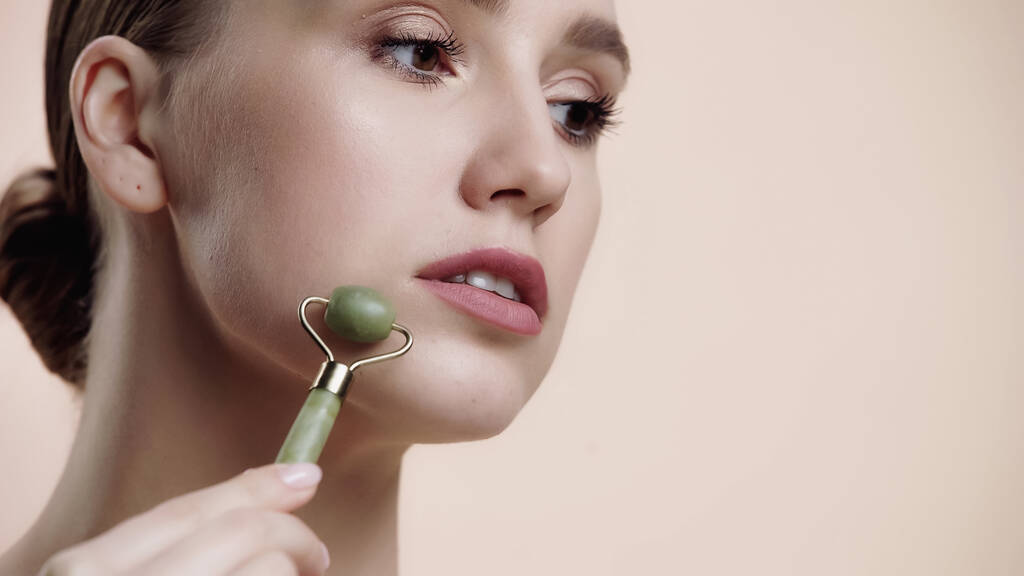 young woman using jade roller while massaging and lifting face isolated on beige  - Fotografie, Obrázek