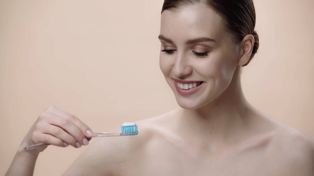 happy woman with bare shoulders holding toothbrush with toothpaste isolated on beige  - Foto, Bild