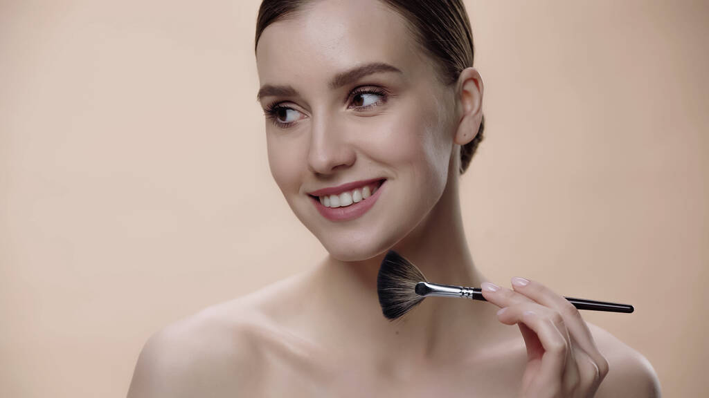 pleased woman with bare shoulders applying face powder while holding cosmetic brush isolated on beige - Fotoğraf, Görsel