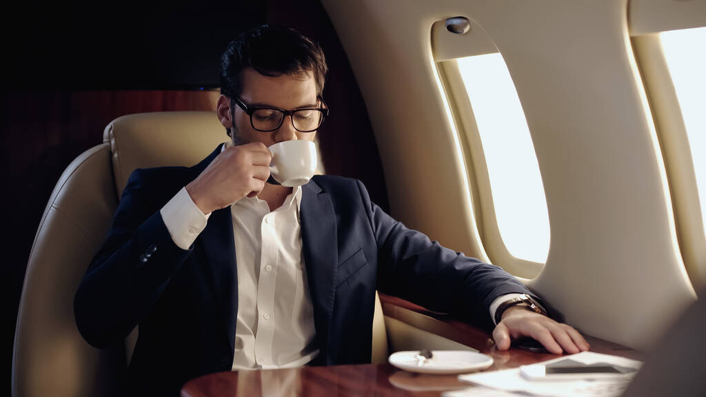 Businessman in eyeglasses drinking coffee near smartphone and papers in private plane  - Photo, Image