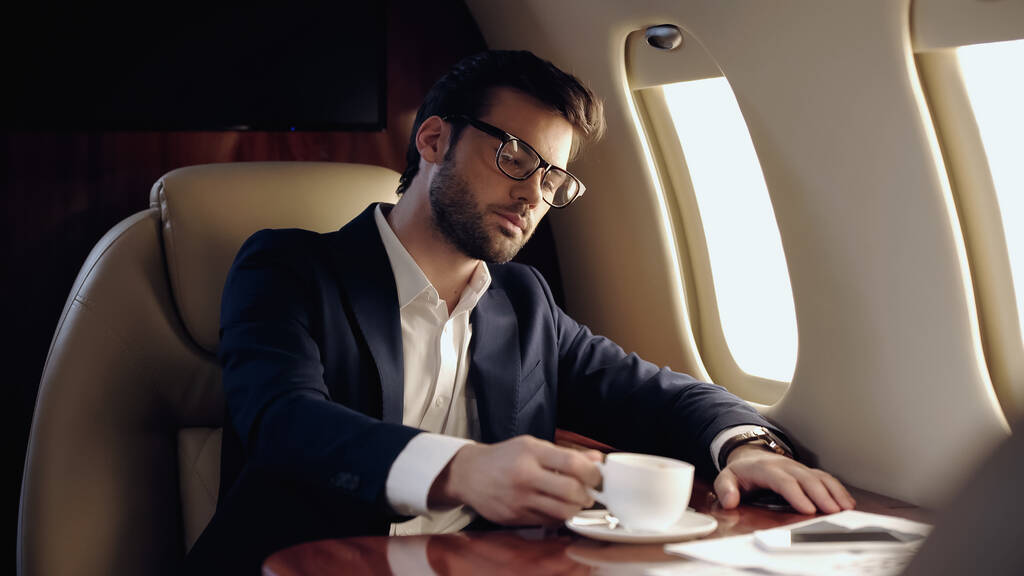Businessman taking cup of coffee near cellphone in private plane  - Photo, image
