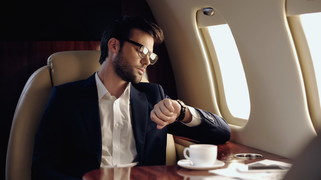 Businessman looking at wristwatch near coffee and cellphone in private plane  - Fotoğraf, Görsel