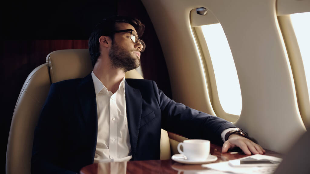 Businessman looking at window near coffee and smartphone in private jet  - 写真・画像