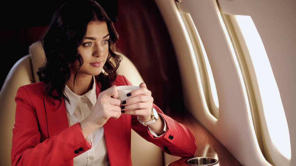 Businesswoman holding cup looking at window in private plane  - Foto, afbeelding