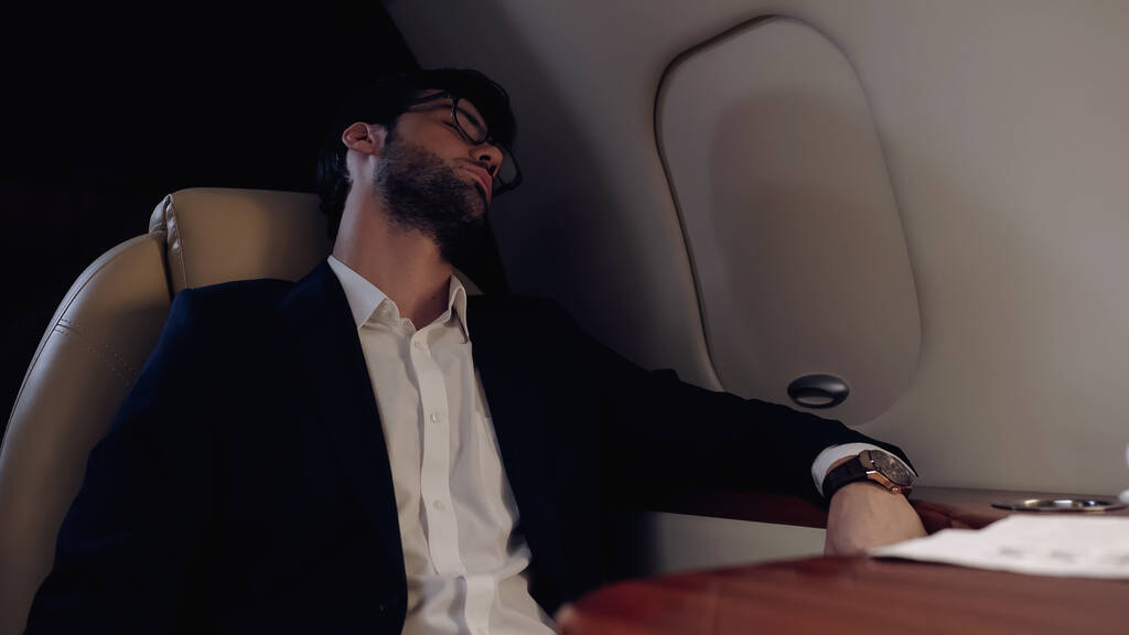 Επιχειρηματίας με γυαλιά ηλίου κοιμάται κοντά σε χαρτιά σε ιδιωτικό αεροπλάνο  - Φωτογραφία, εικόνα