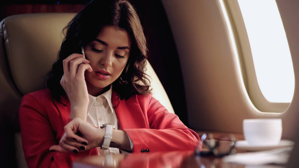 Businesswoman talking on smartphone and looking at wristwatch near cup in private plane  - Фото, изображение