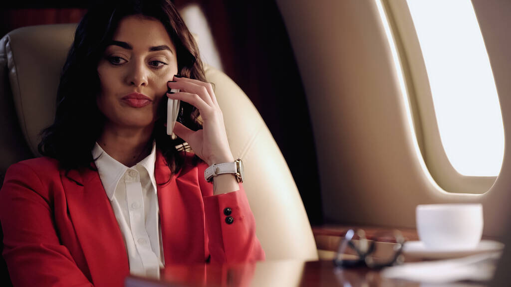 Businesswoman talking on smartphone near blurred cup in private plane  - Zdjęcie, obraz