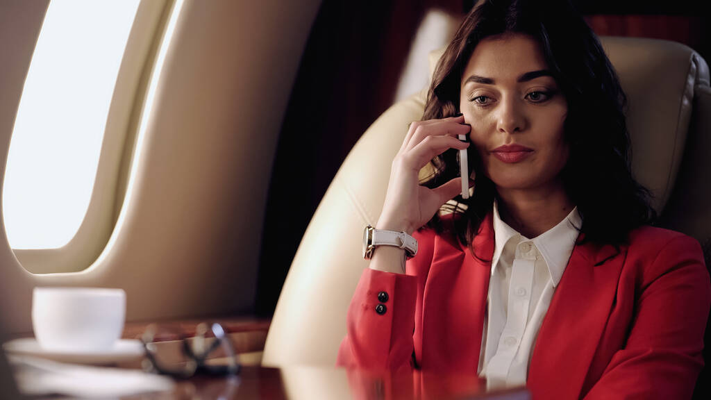 Femme d'affaires parlant sur un téléphone portable près de tasse floue et des lunettes dans un avion privé  - Photo, image
