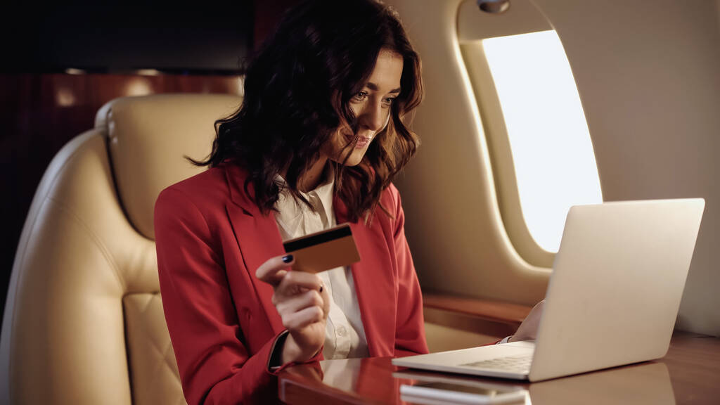 Femme d'affaires souriante utilisant une carte de crédit et un ordinateur portable dans un avion privé  - Photo, image
