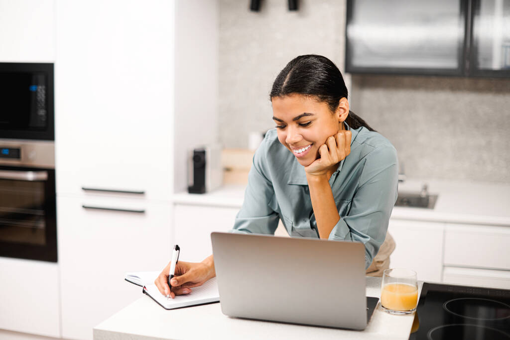 Positiv intelligente junge Studentin mit gemischter Rasse lächelt, während sie am Tisch mit Laptop in der Küche steht, von zu Hause aus lernt und Notizen zum Notizbuch macht und sich ein Online-Webinar ansieht - Foto, Bild
