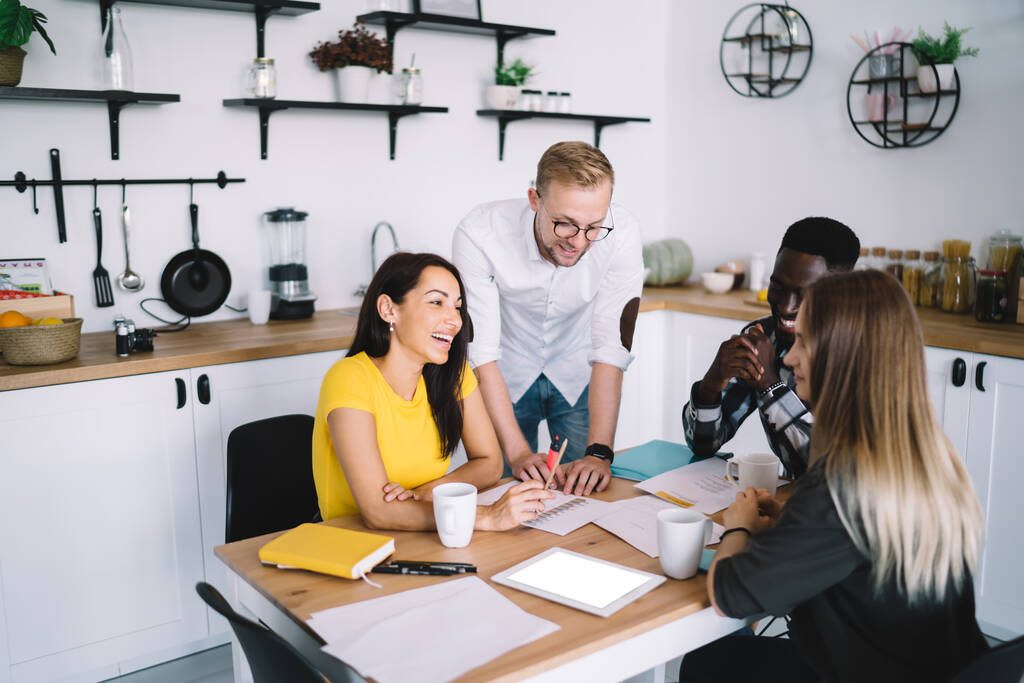 High angle of happy multiracial colleagues in casual outfits discussing business plan while working with papers at wooden table in kitchen - Foto, afbeelding