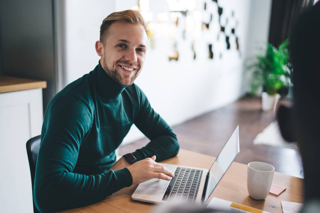 Young handsome businessman in green turtleneck sitting at table with laptop and looking at camera with smile while working on business project in creative workspace shared with other entrepreneurs - Foto, Bild